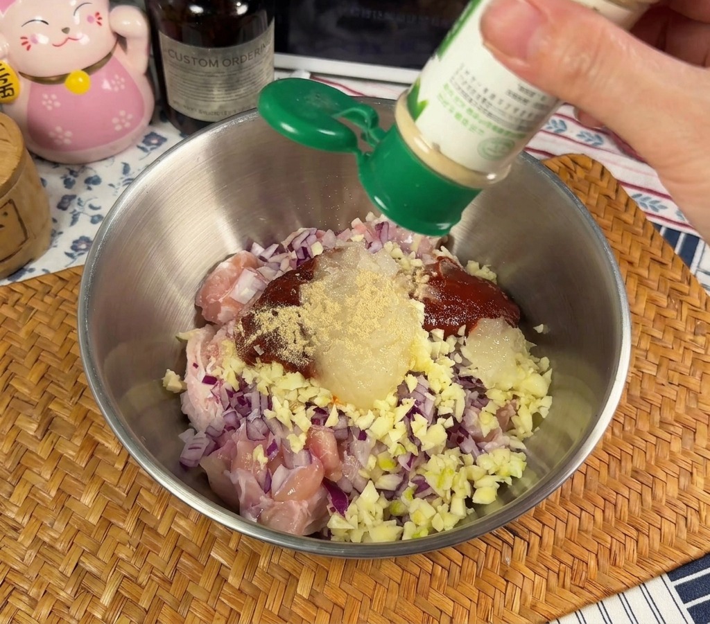 Una persona spolvera del pepe bianco in polvere da un barattolino verde in una ciotola di metallo contenente pollo crudo e cipolle rosse tritate.