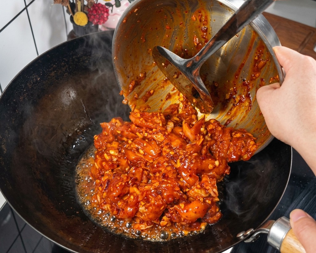 Pezzi di pollo marinati in una salsa rossa intensa vengono versati da una ciotola di metallo in un wok nero ben caldo e fumante.