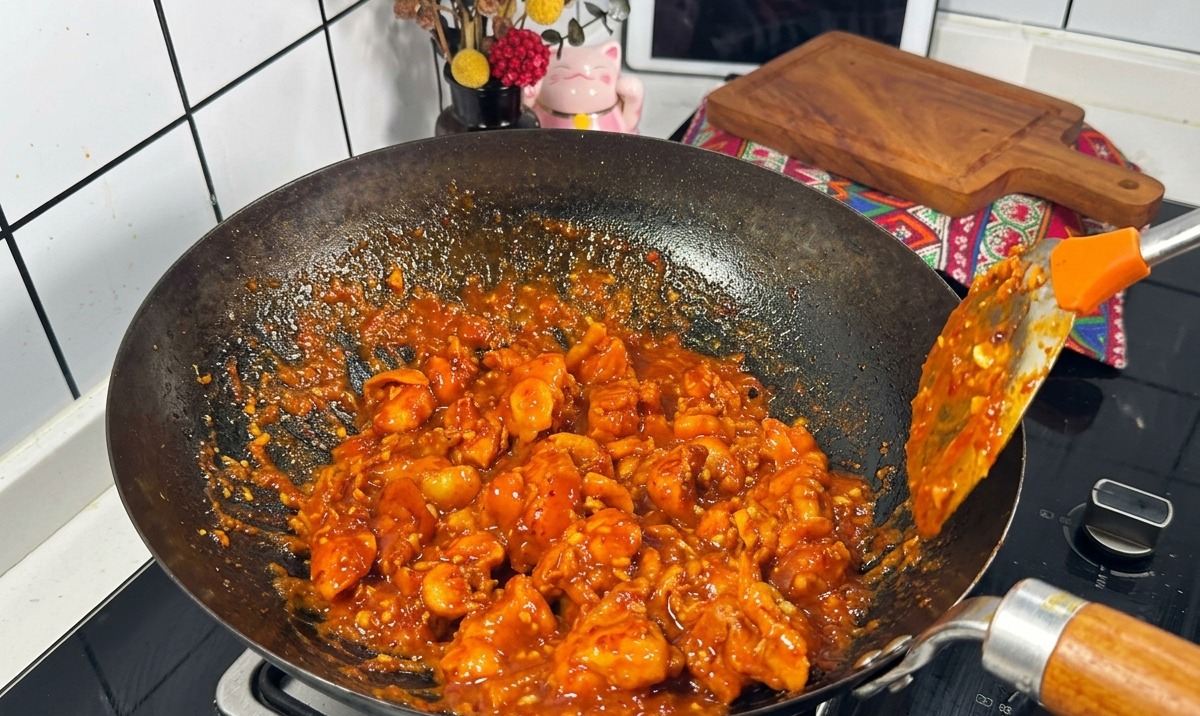 Pezzi di pollo avvolti da una densa salsa rossa che sobbolle vengono saltati in un wok nero con una spatola di metallo.