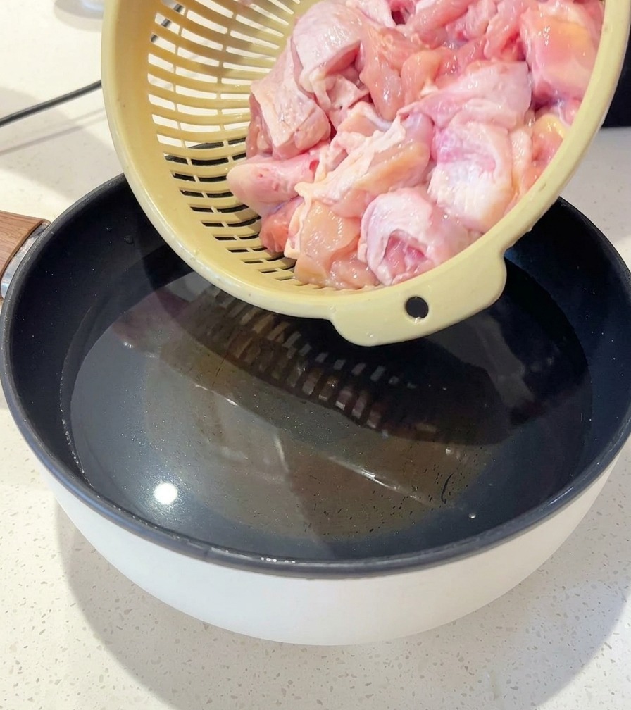 Una persona che versa pezzi di pollo crudo da un colino in una pentola dacqua con zenzero a fette e cipollotti.