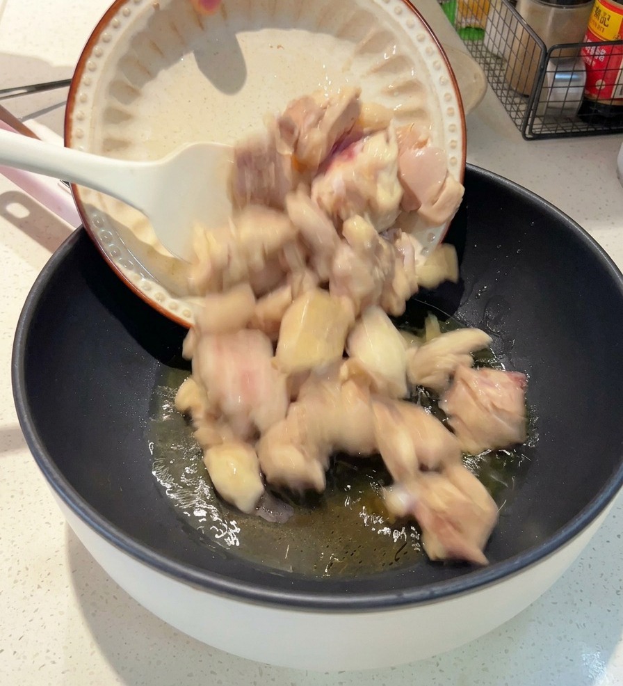 Un piatto bianco da cui vengono versati pezzi di pollo sbollentati in una padella scura con olio sfrigolante.