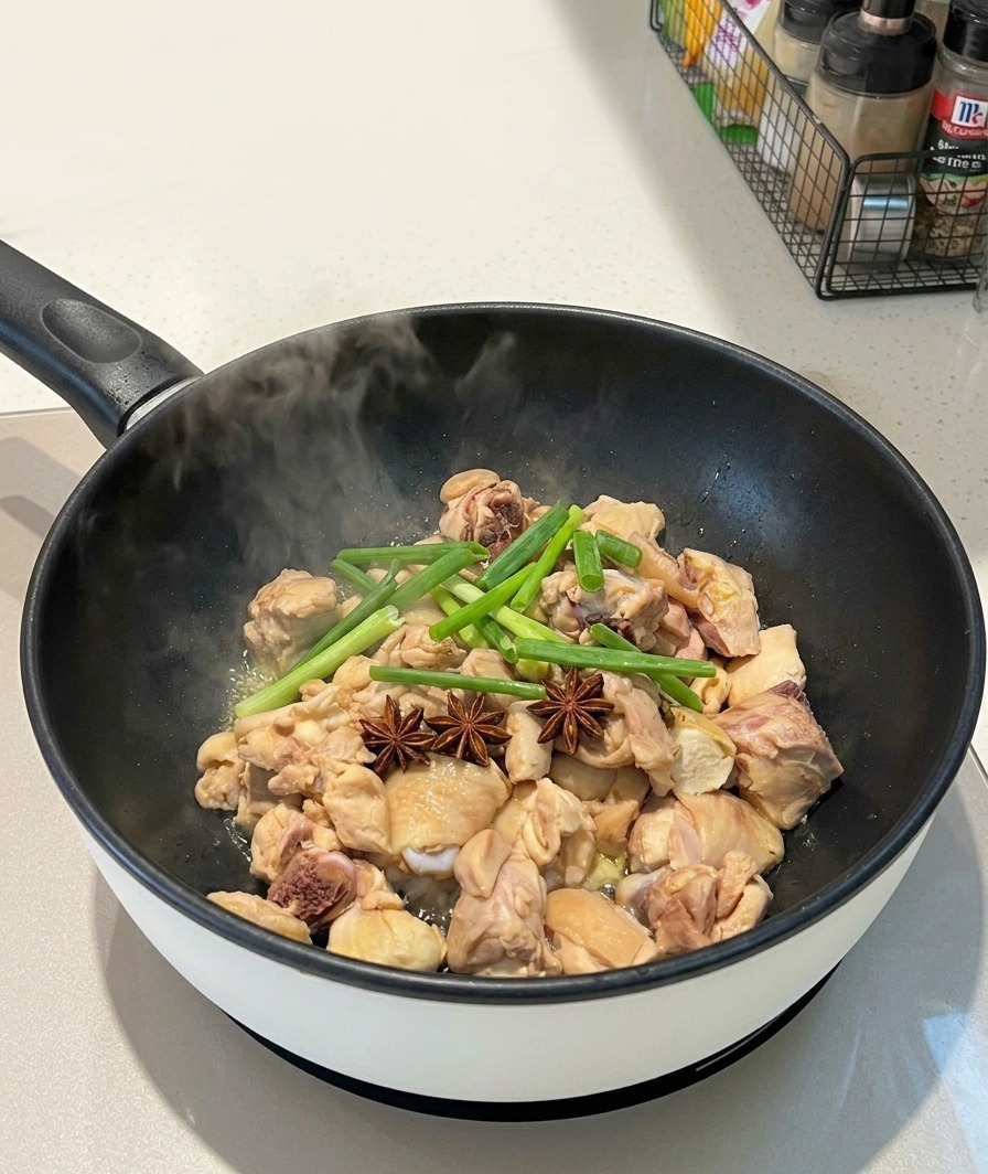 Anice stellato intero e gambi di cipollotto verde adagiati sopra i pezzi di pollo in una padella fumante.