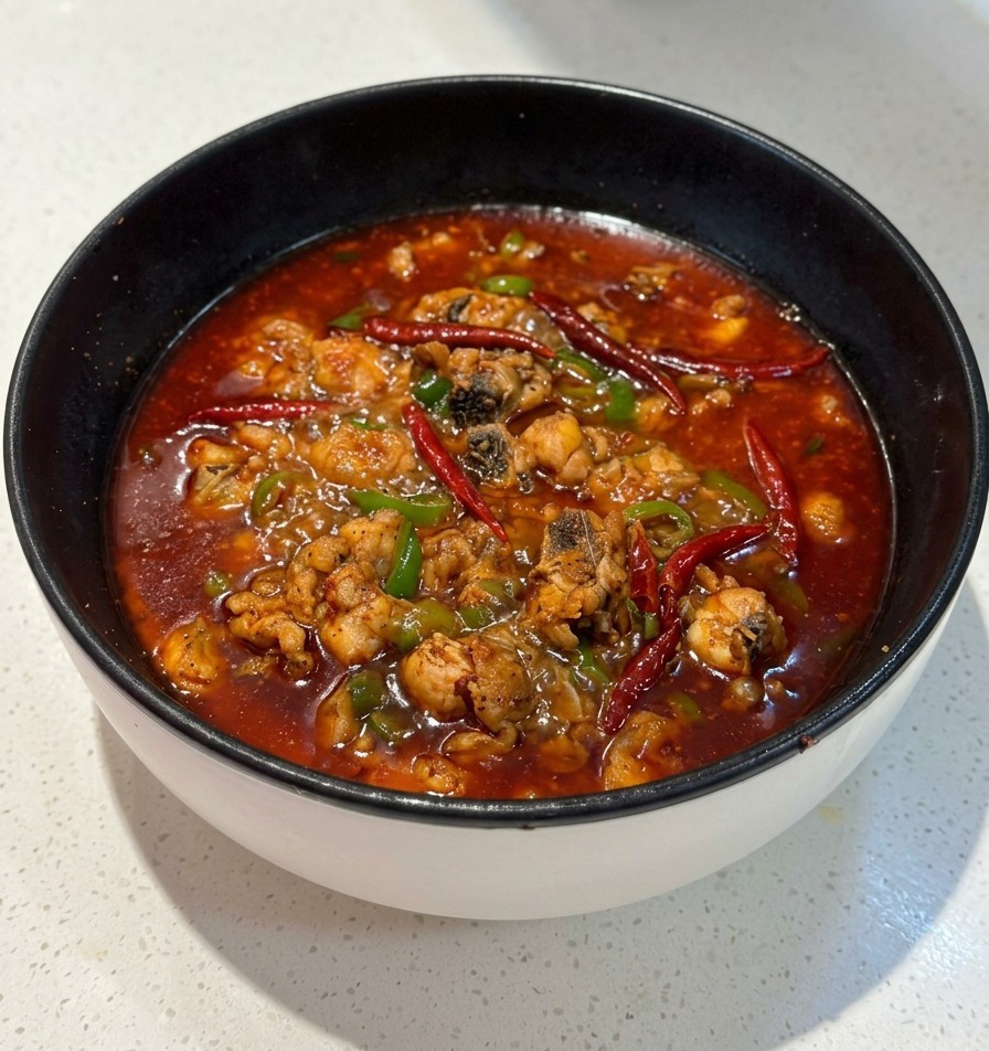 Una padella nera piena di un brodo rosso piccante e ribollente che contiene pezzi di pollo e segmenti di cipollotto verde.