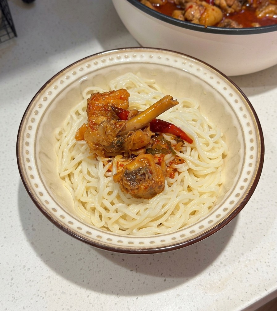 Una ciotola di ceramica con noodle bianchi sormontati da pollo piccante e salsa al peperoncino rosso, con la padella di cottura visibile sullo sfondo.