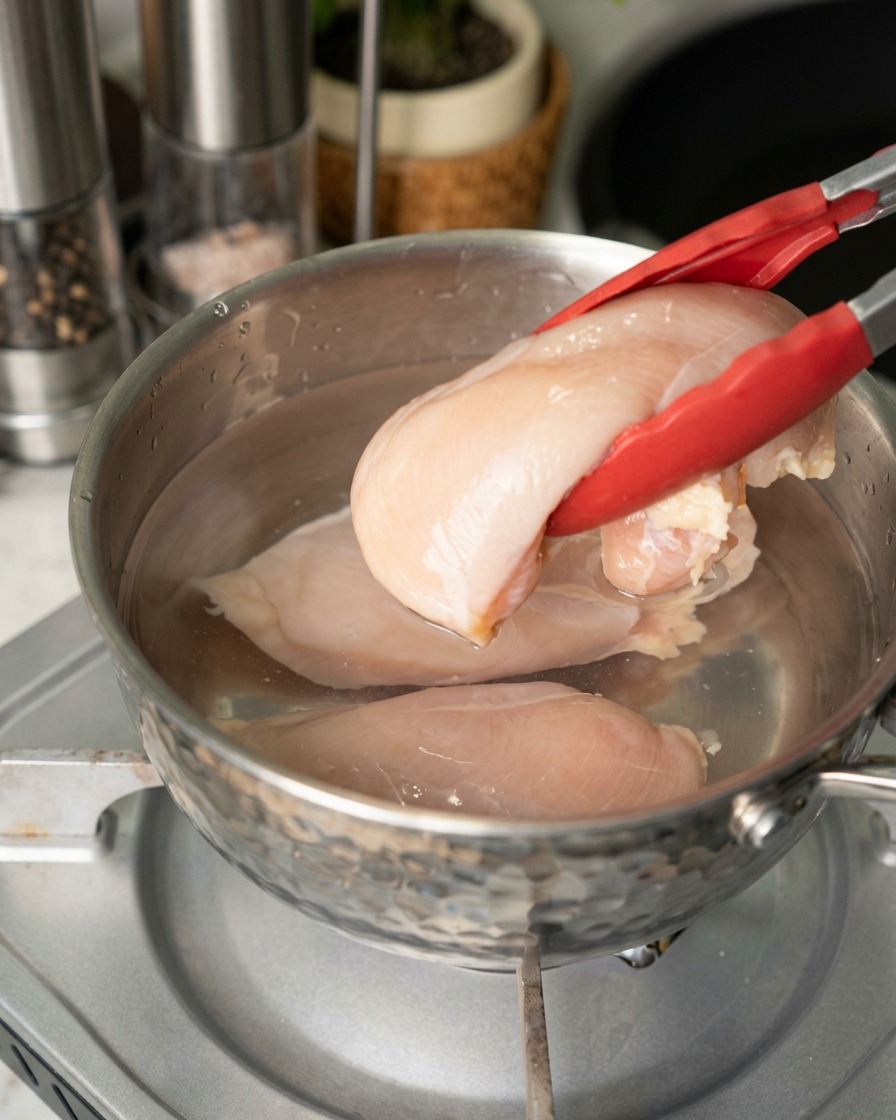 Una persona usa delle pinze con punta rossa per immergere petti di pollo crudi e rosati in una pentola dacciaio con acqua.