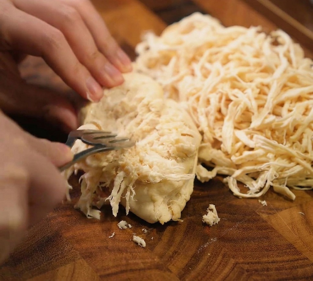 Due forchette di metallo vengono usate per sfilacciare un petto di pollo intero cotto in striscioline sottili su un tagliere di legno scuro.