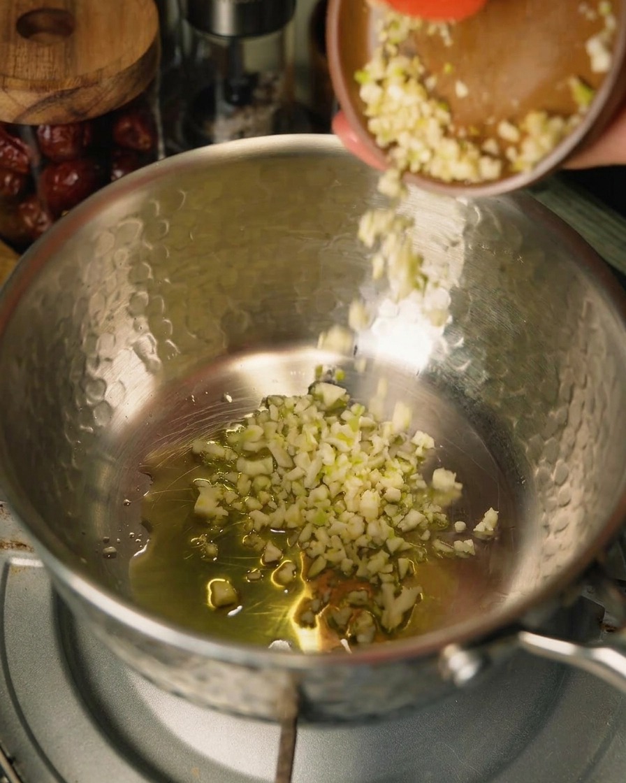 Aglio tritato viene versato da una ciotolina marrone in una pentola con olio e cipolle, si nota il bordo di una spatola rossa.
