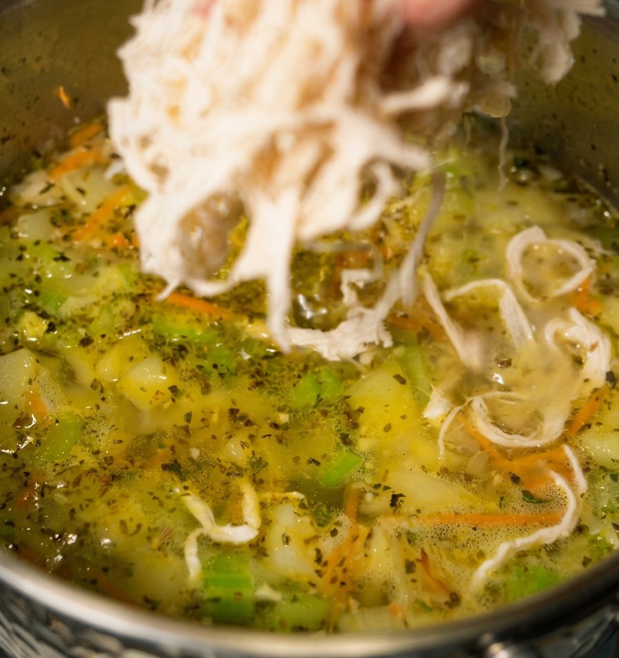 Una mano lascia cadere straccetti di pollo bianco in una pentola di zuppa di verdure che sobbolle, con carote e sedano visibili.