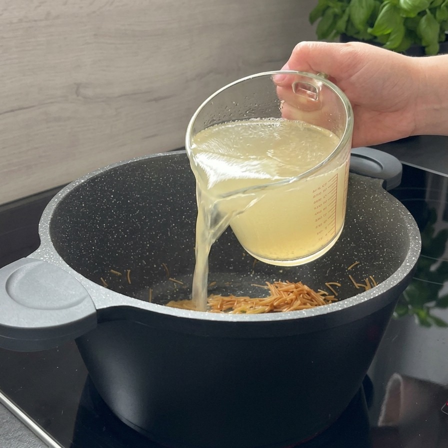 Brodo di pollo limpido versato da un misurino di vetro in una pentola nera con la pastina tostata.