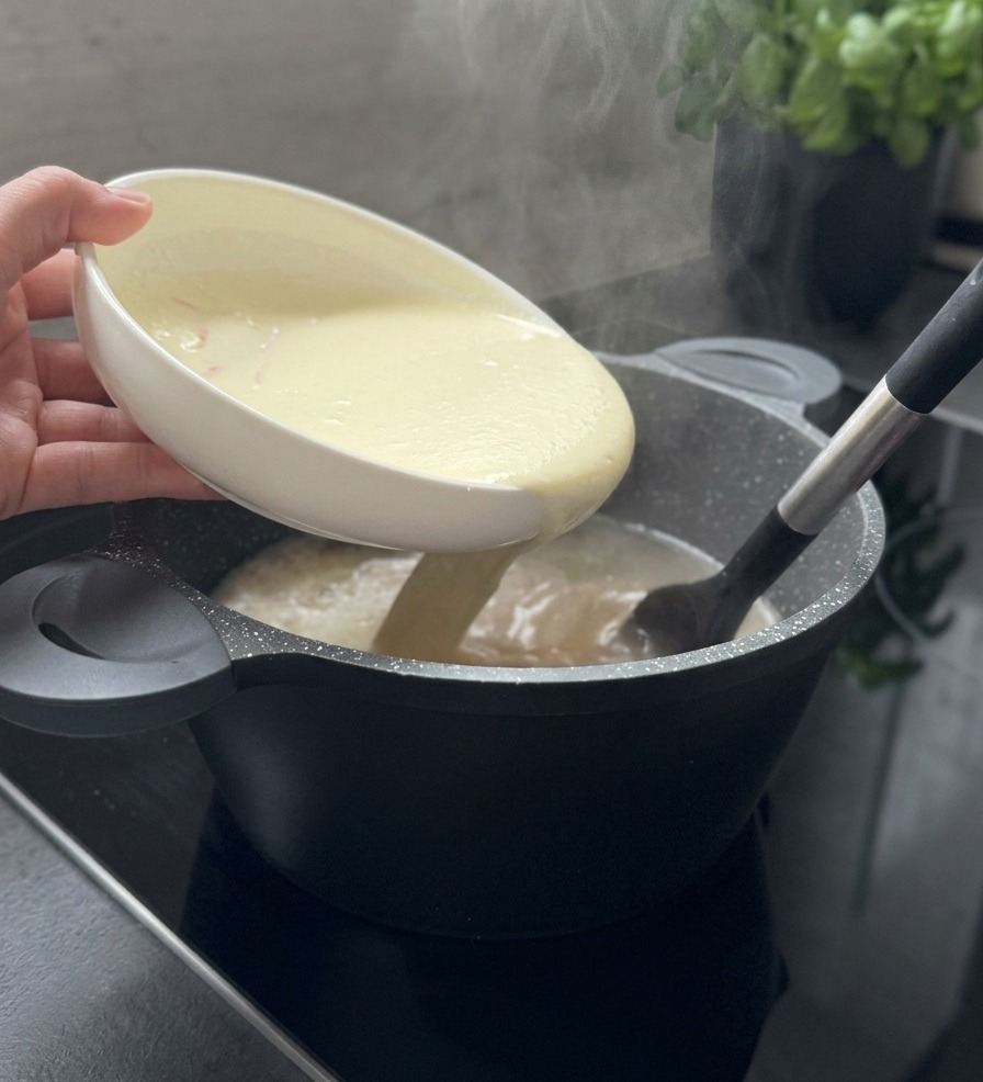 Un composto cremoso e chiaro viene versato da una ciotola bianca in una pentola di zuppa fumante mentre si mescola con una spatola nera.