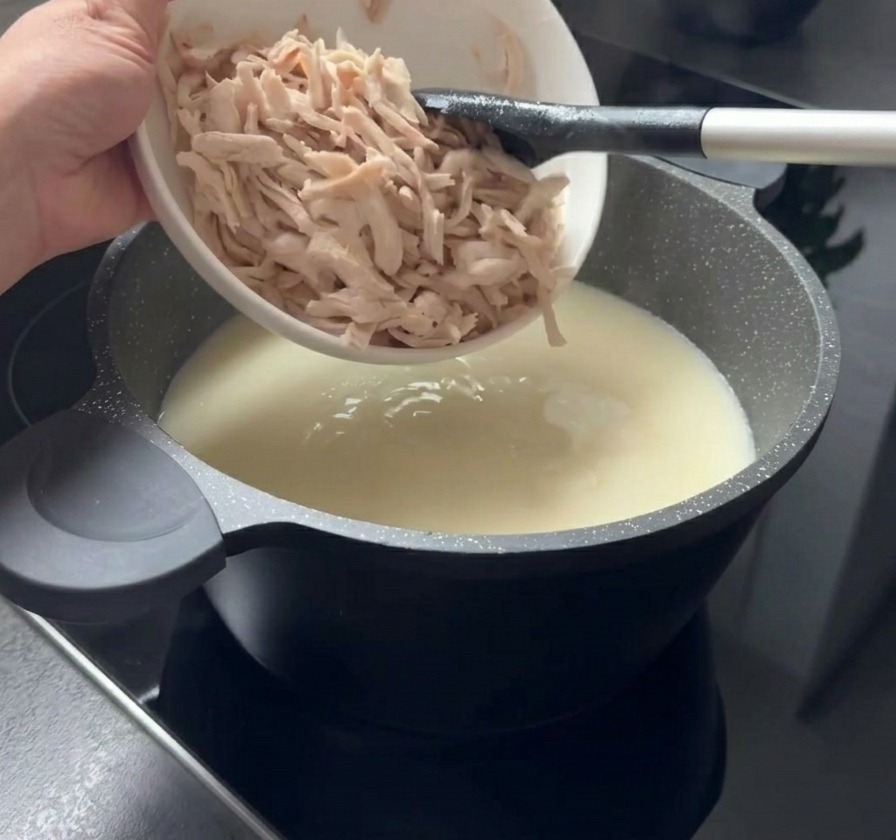 Pezzi sfilacciati di pollo cotto aggiunti da una ciotola bianca in una pentola di zuppa cremosa e chiara.