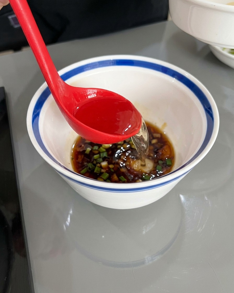 Un mestolo rosso versa acqua calda e limpida in una ciotola bianca contenente salsa di soia scura, olio e aromi tritati.