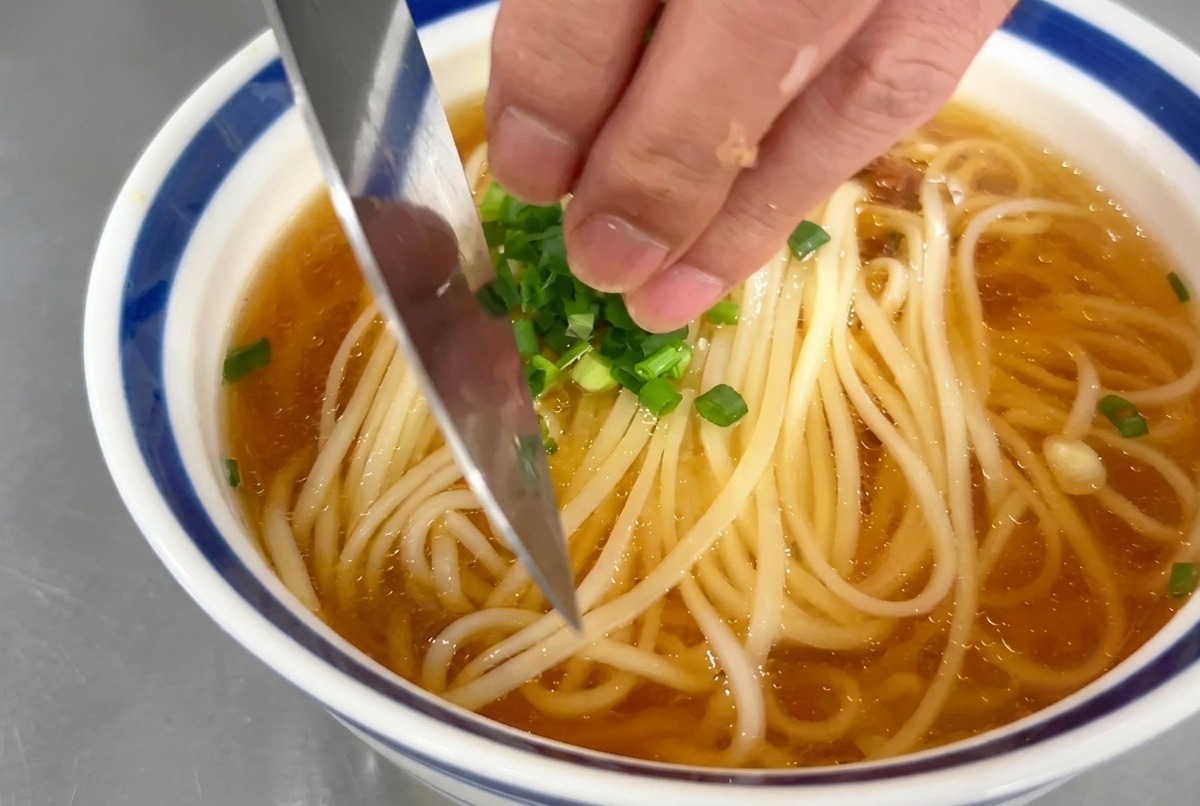 Una mano spolvera rondelle di cipollotto verde su una ciotola di zuppa di noodle.
