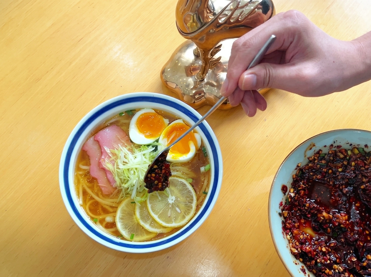 Una persona usa un lungo cucchiaio di metallo per adagiare una punta di pasta di olio al peperoncino rosso scuro sopra un ciuffo di ravanello bianco grattugiato in una ciotola di noodle.