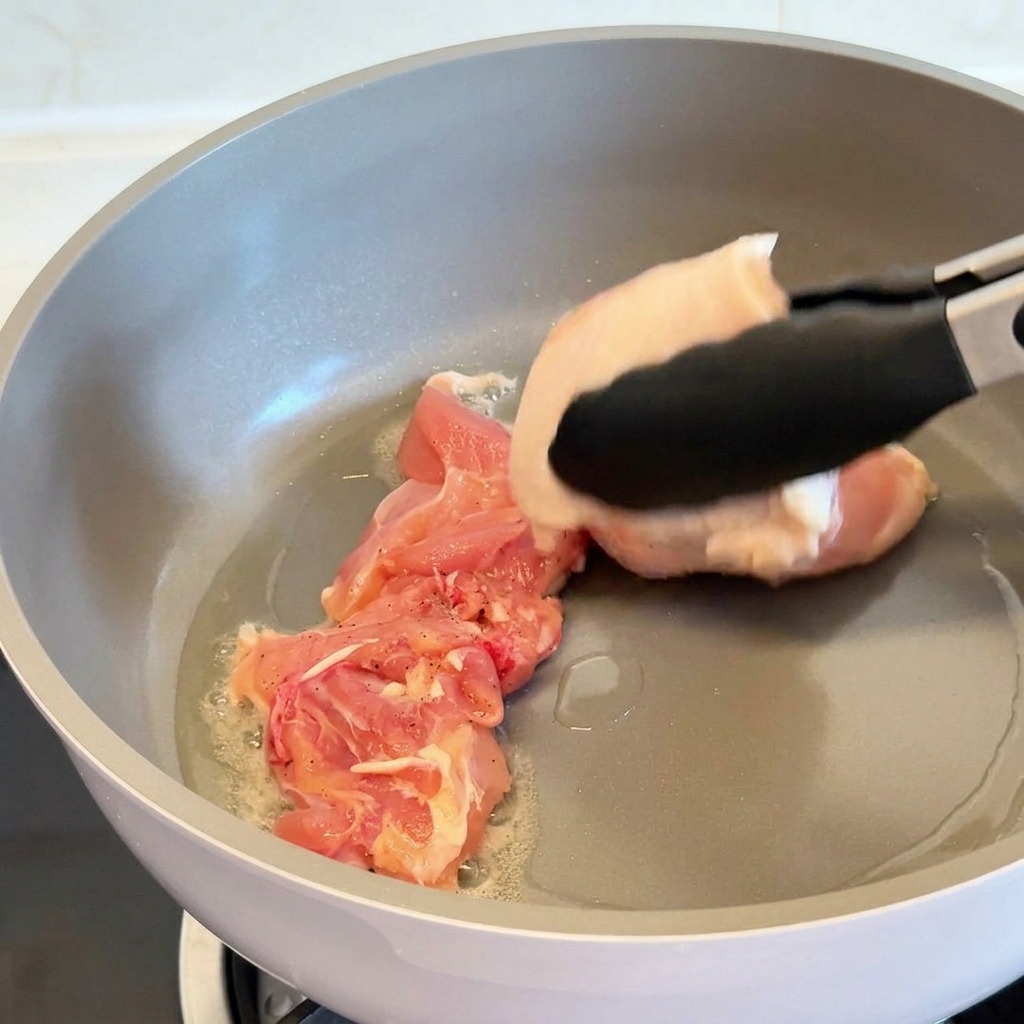 Pinze da cucina che adagiano una sovracoscia di pollo cruda con la pelle rivolta verso il basso in una padella antiaderente grigia con olio caldo.