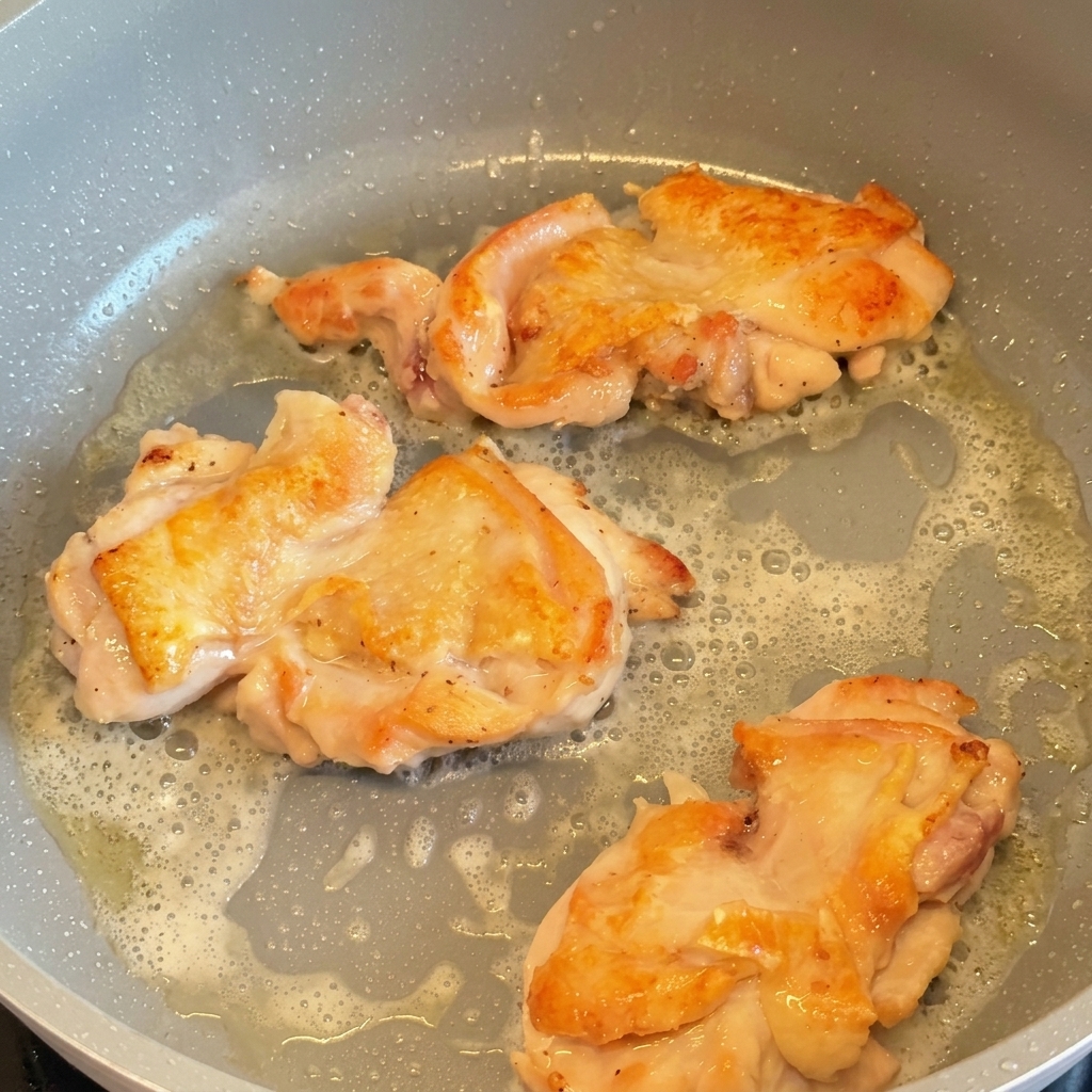 Tre sovracosce di pollo che friggono in padella, girate per mostrare la pelle rosolata e dorata.