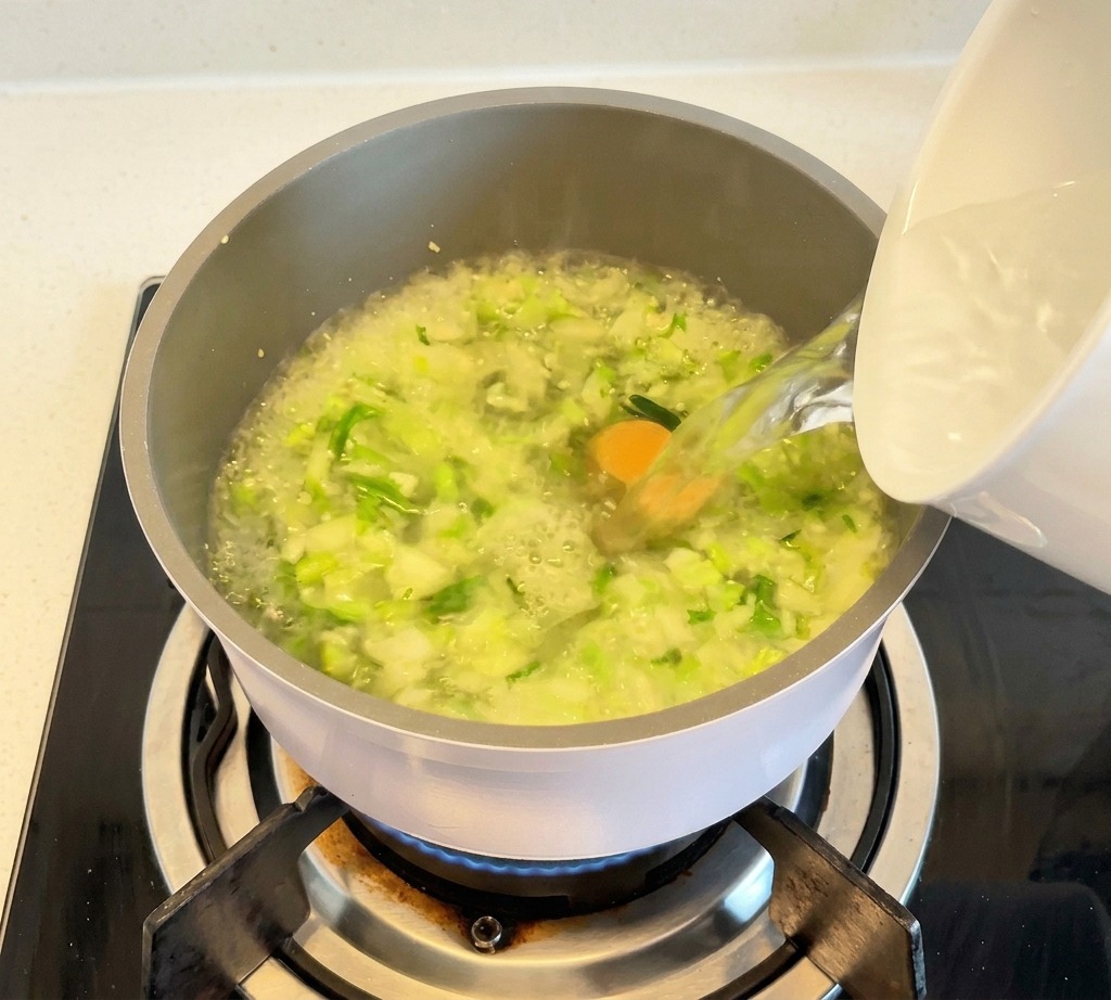 Acqua bollente versata da una ciotola bianca in una pentola colma di verdure per formare un brodo limpido.
