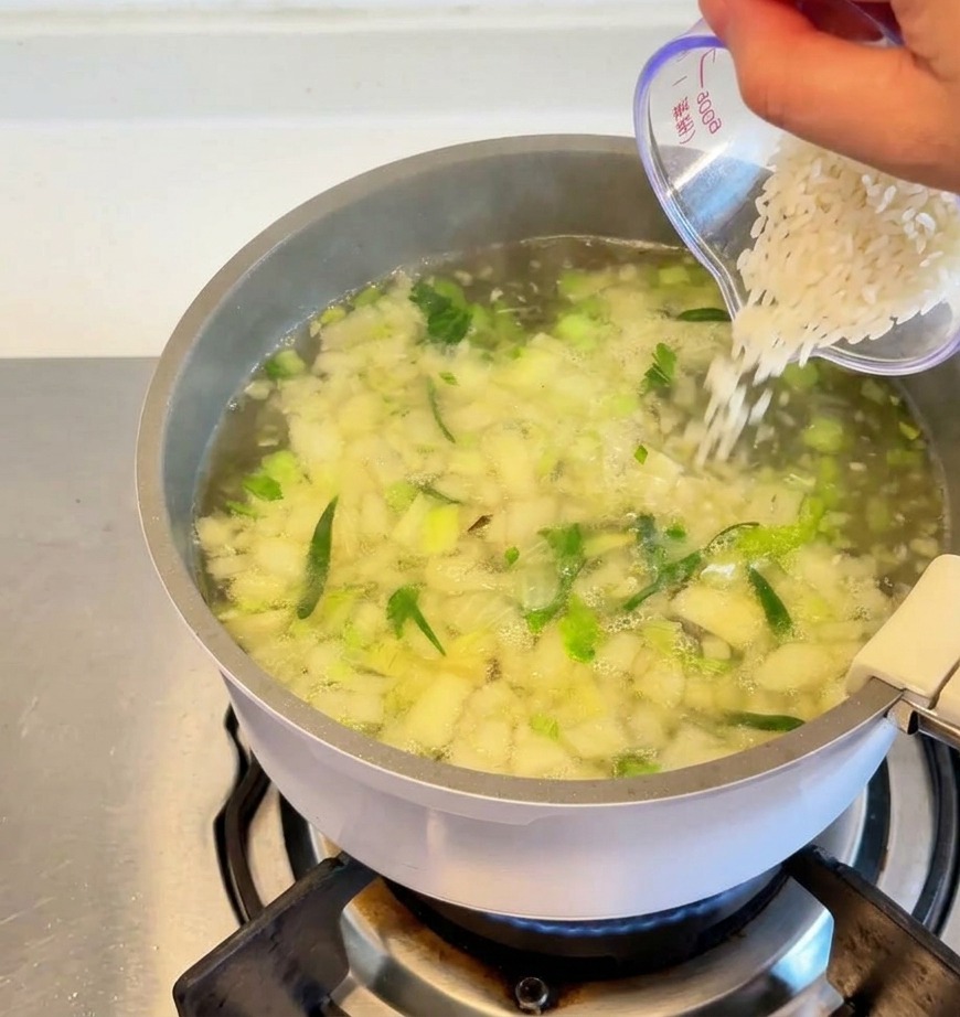 Riso bianco versato da un misurino trasparente in una pentola di zuppa che sobbolle con verdure ed erbe visibili.