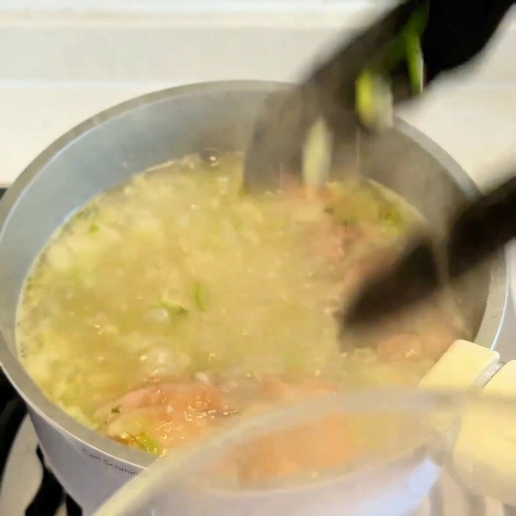 Una pinza da cucina che solleva una sovracoscia di pollo cotta e dorata da una pentola di brodo in ebollizione.