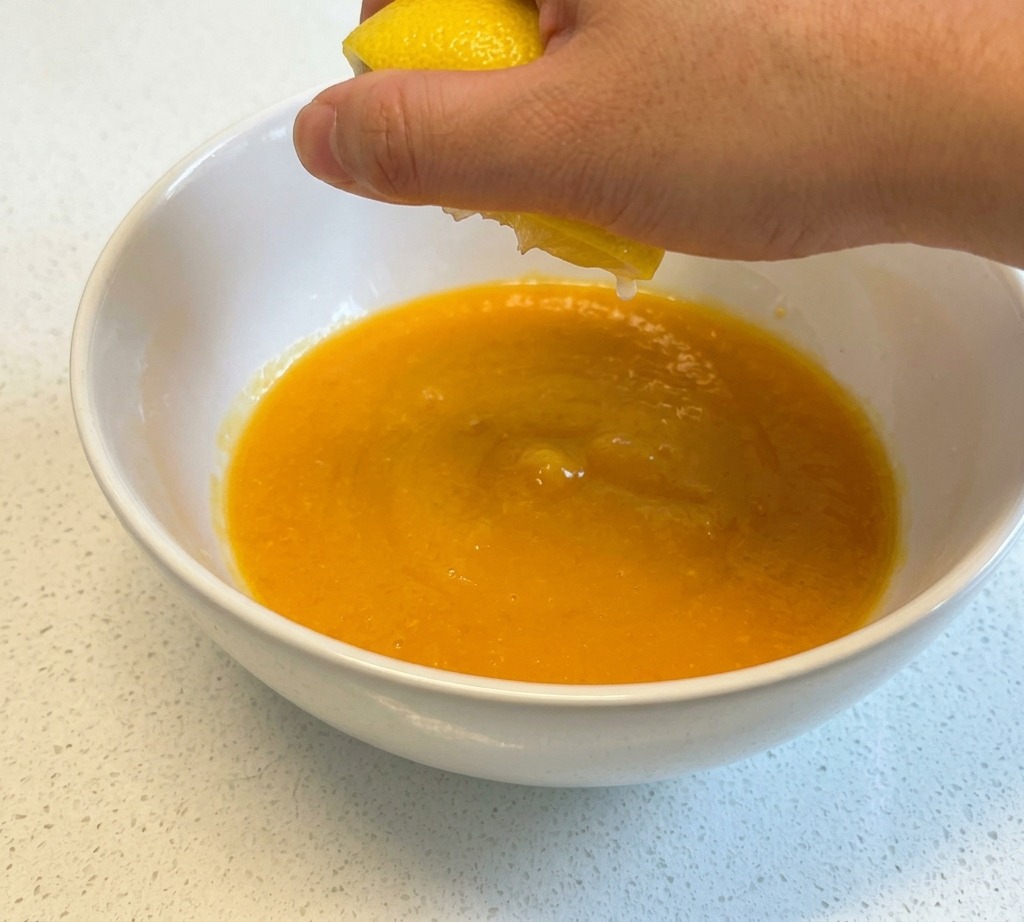 Una mano che spreme il succo da mezzo limone giallo in una ciotola di ceramica bianca contenente uova sbattute.