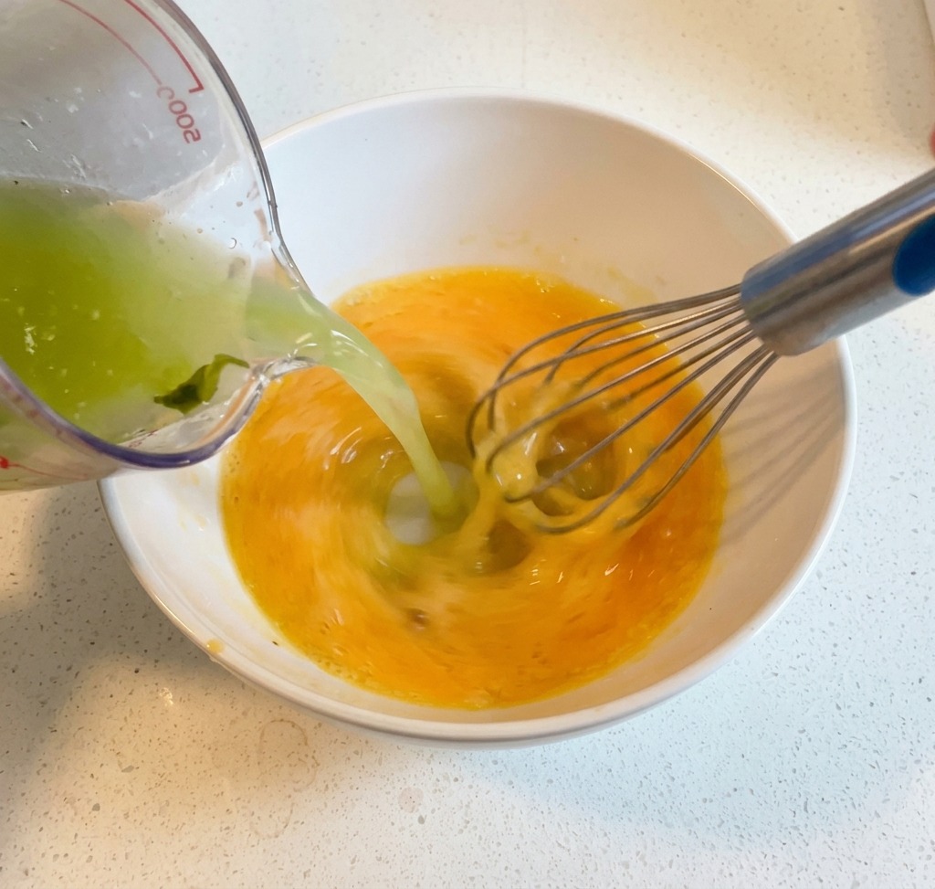 Brodo caldo versato da un mestolo in una ciotola con uova sbattute e limone mentre viene mescolato energicamente con una frusta a mano.