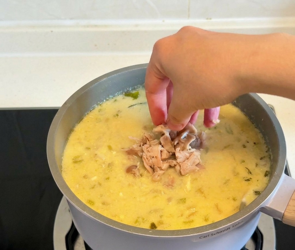 Mani che tuffano pezzi di pollo cotto e sfilacciato in una pentola di zuppa densa e gialla.