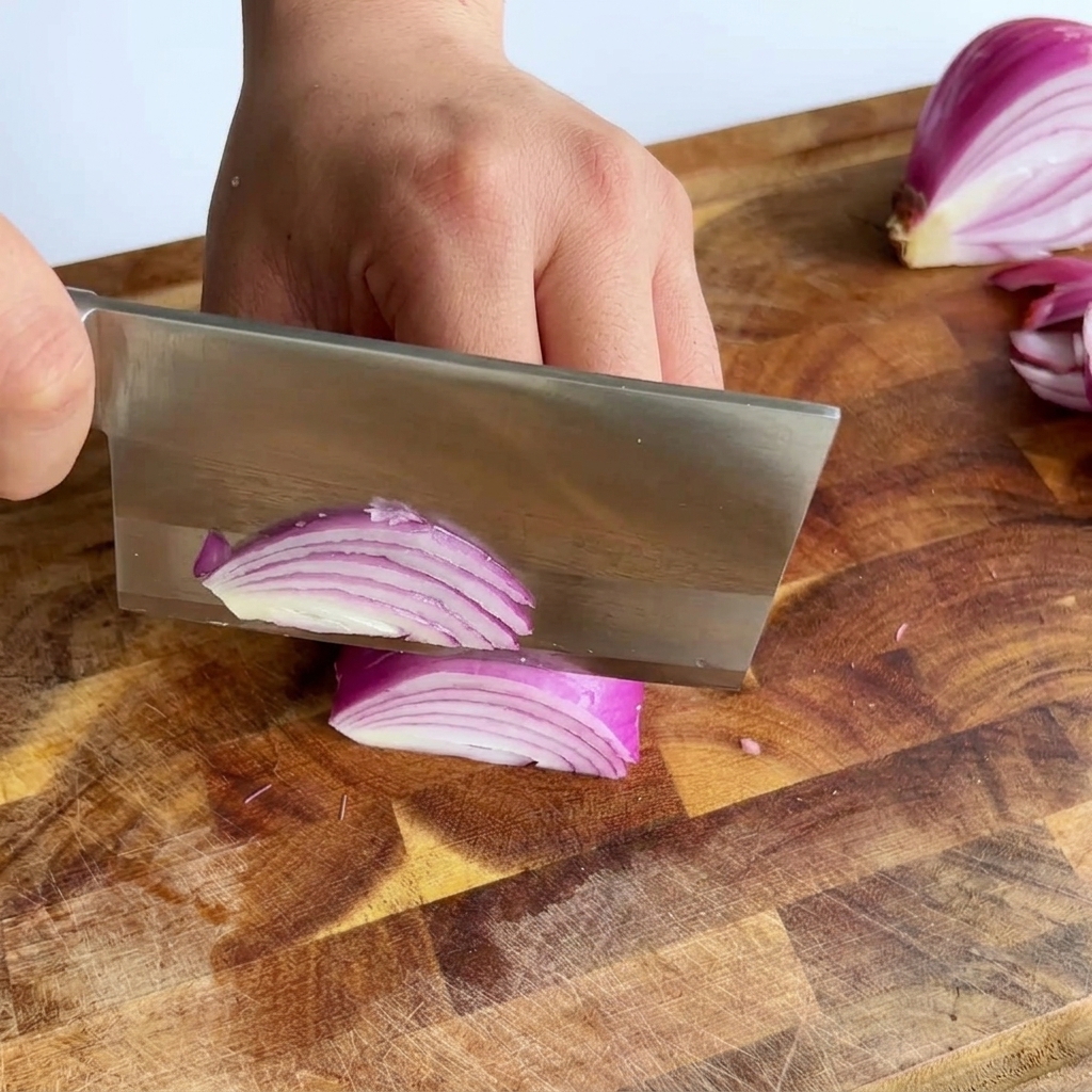 Un grande coltello che affetta una cipolla rossa creando sottili strisce viola su un tagliere di legno.