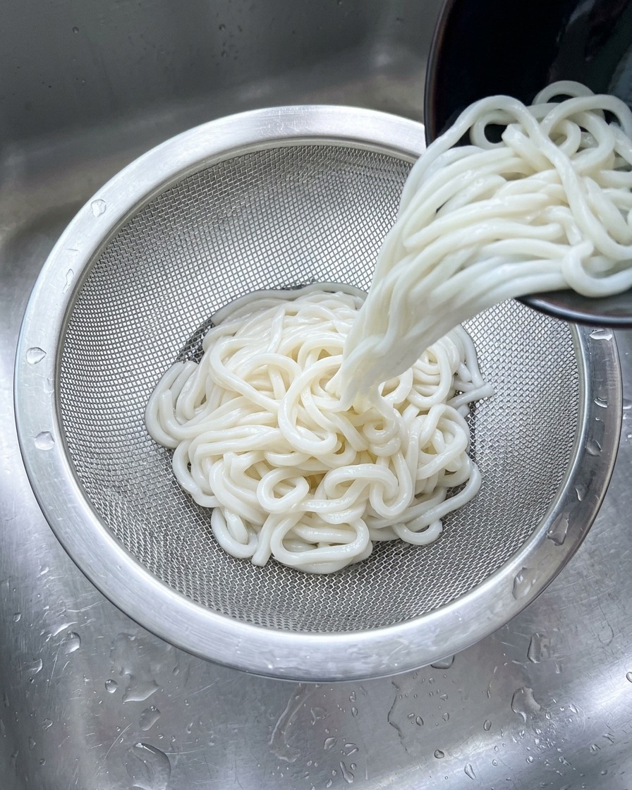 Udon bianchi versati in un colino dacciaio inossidabile sotto un getto dacqua fredda nel lavandino.