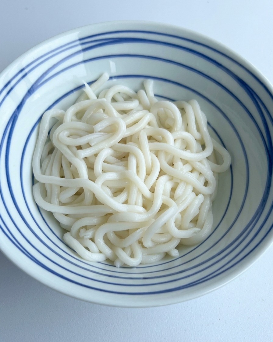 Salsa di soia scura e condimenti versati sopra un mucchio di udon bianchi in una ciotola a righe blu.