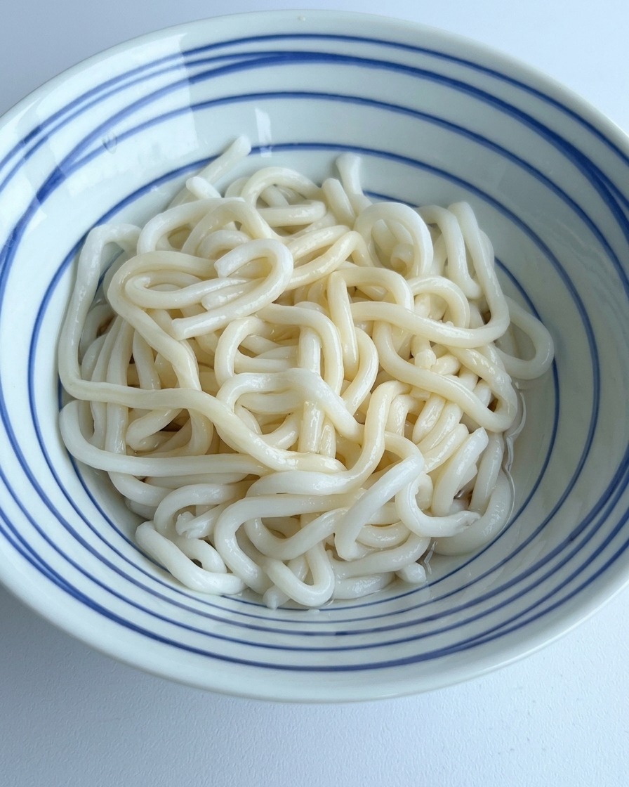 Acqua limpida versata in una ciotola di udon che si mescola con i condimenti scuri sul fondo.