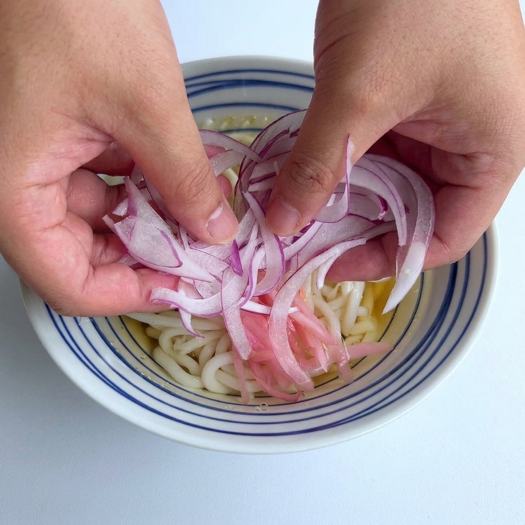 Due mani che adagiano un mucchietto di cipolle rosse affettate sottili al centro di una ciotola di udon.