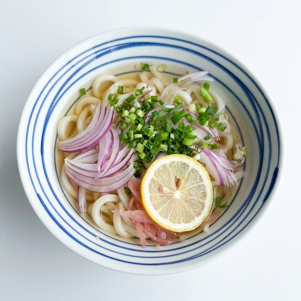 Vista dallalto di una ciotola di udon in brodo limpido, conditi con cipolle rosse e cipollotti, con una fetta di limone giallo al centro.