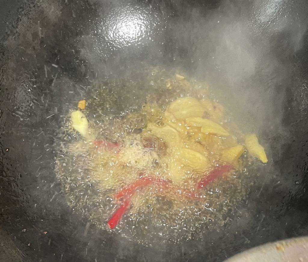 Fette di zenzero e peperoncini rossi secchi che sfrigolano nellolio caldo dentro un wok scuro, rilasciando vapore e profumi.