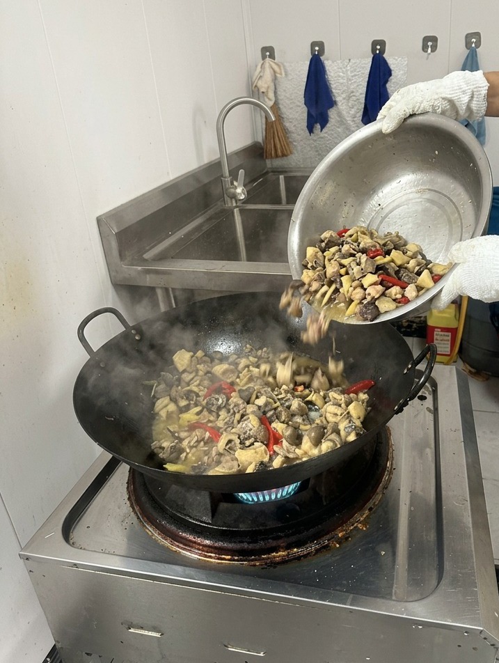 Una persona che versa acqua limpida bollente da un contenitore in un wok fumante con pollo rosolato e spezie.