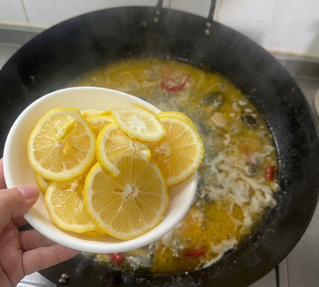 Una mano che tient una ciotola bianca con sottili fette di limone sopra un wok in cui bolle un brodo di pollo dorato.