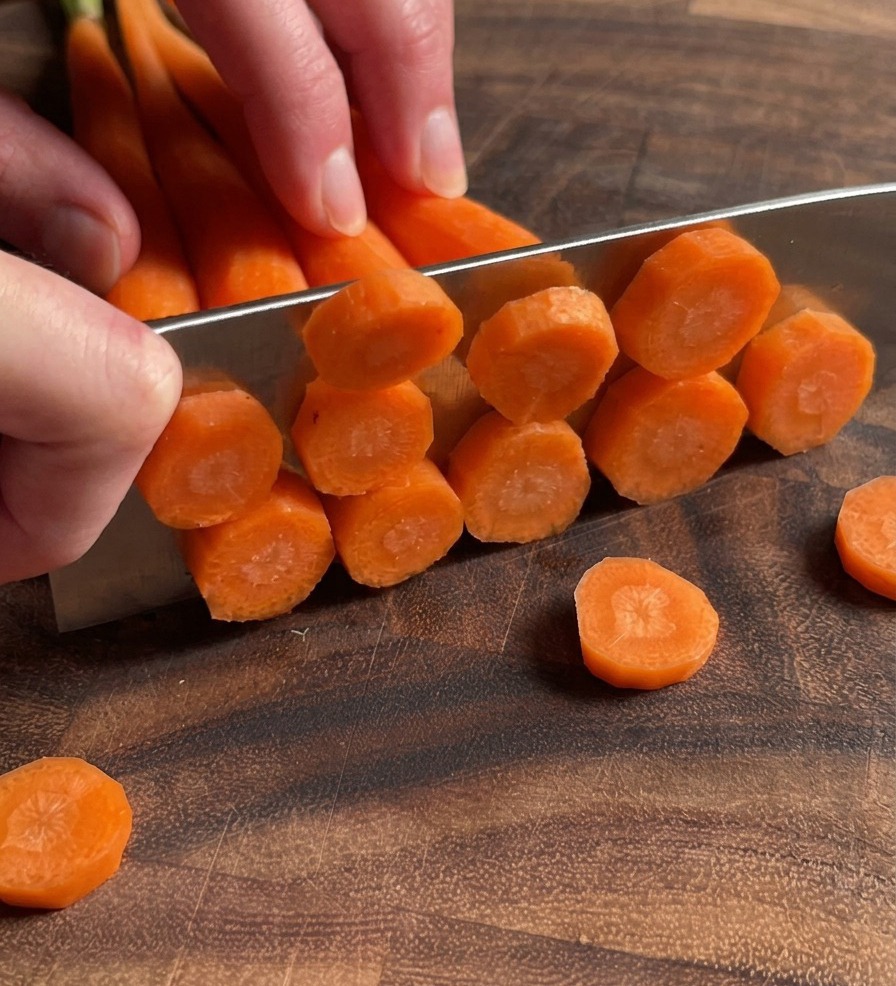 Un primo piano di mani che tagliano a rondelle sottili diverse carote su un tagliere di legno scuro.