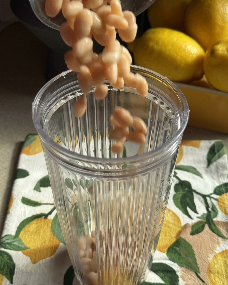 Fagioli bianchi cotti che vengono versati nel boccale trasparente di un frullatore appoggiato su un canovaccio con motivi di limoni.