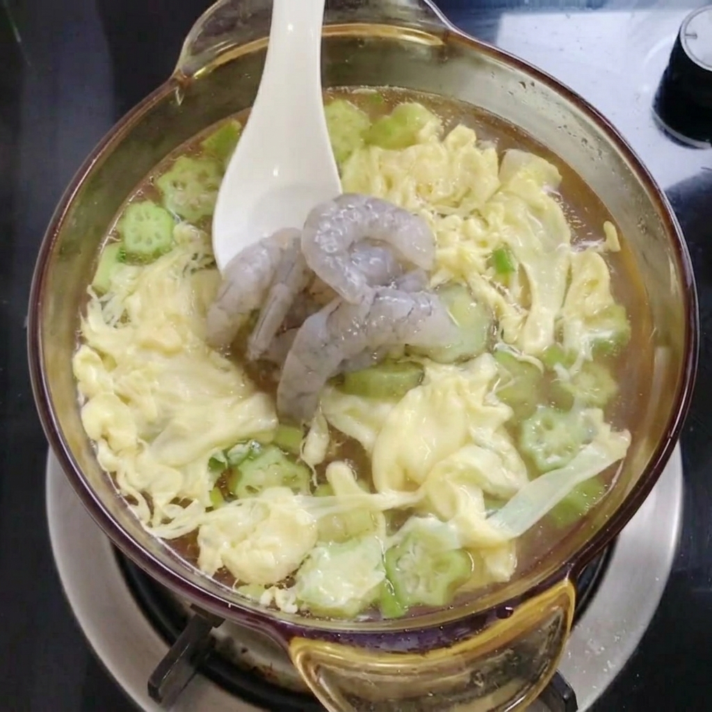 Un cucchiaio bianco che adagia diversi gamberi grigi crudi in una pentola di vetro piena di filamenti duovo gialli e fettine di okra verde nel brodo.