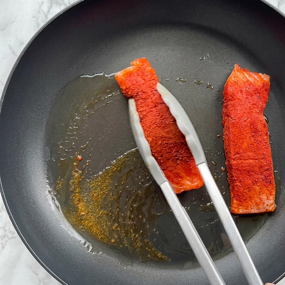 Un filetto di salmone speziato viene scottato in una padella ben calda con olio doliva bollente, tenuto con delle pinze metalliche.