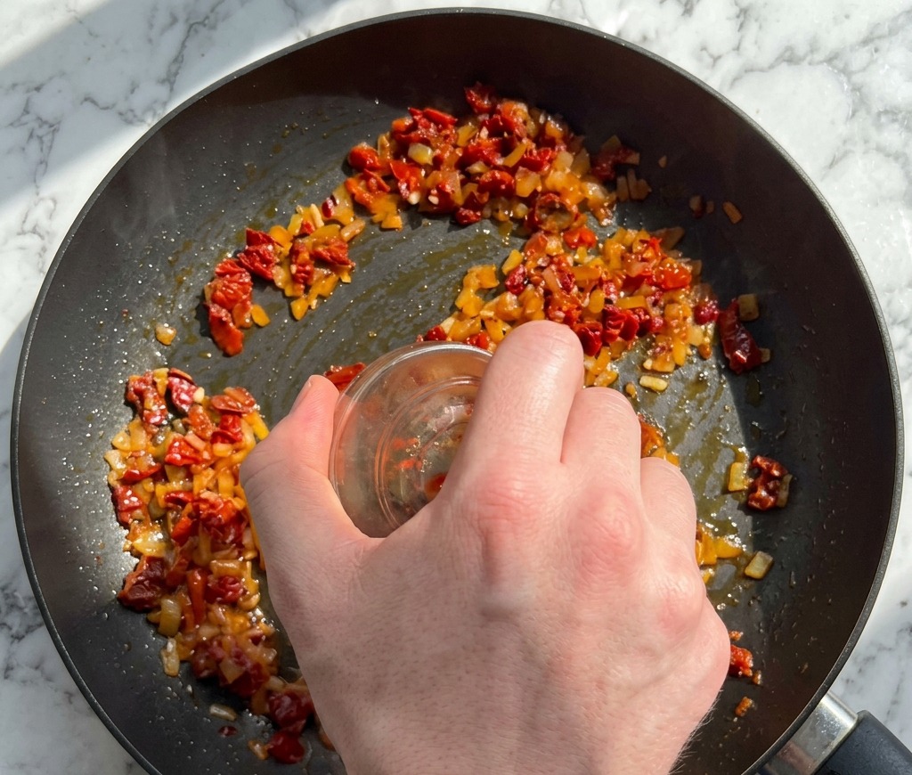 Una mano versa pomodori secchi tritati e fiocchi di peperoncino da una ciotolina di vetro in una padella con gli aromi.