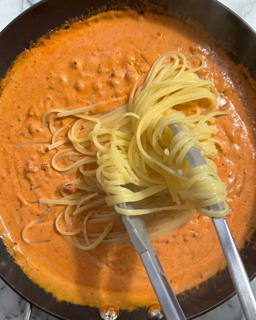 Un nido di pasta cotta viene sollevato con le pinze e adagiato in una padella piena di una ricca salsa arancione e cremosa.