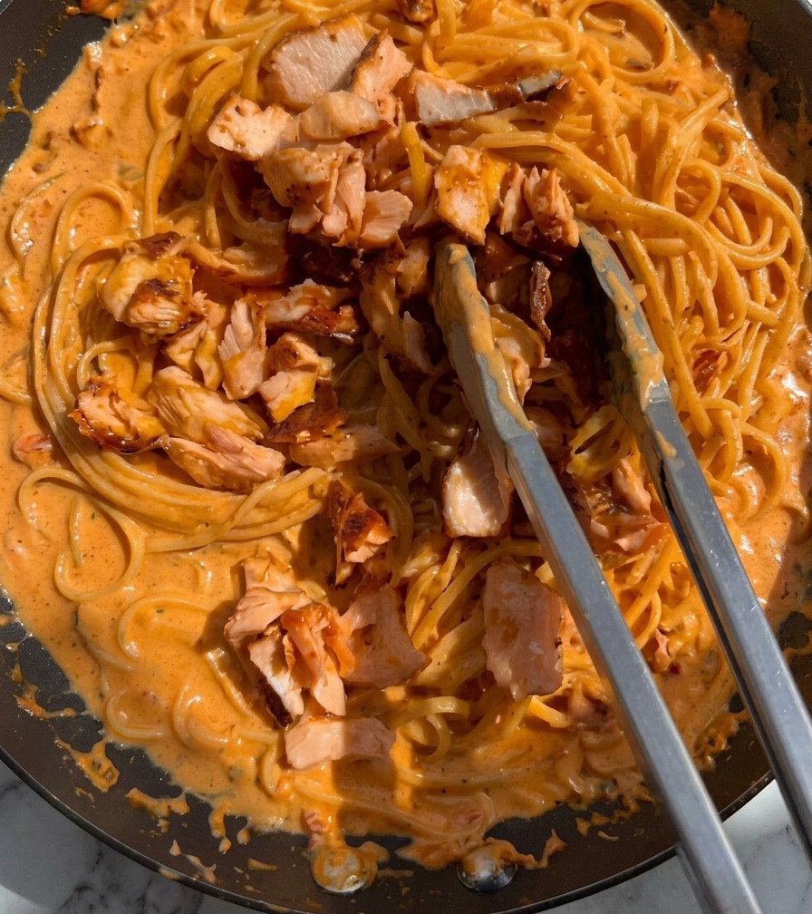 Pezzi di salmone rosa cotto vengono aggiunti a una padella di pasta cremosa usando delle pinze metalliche.
