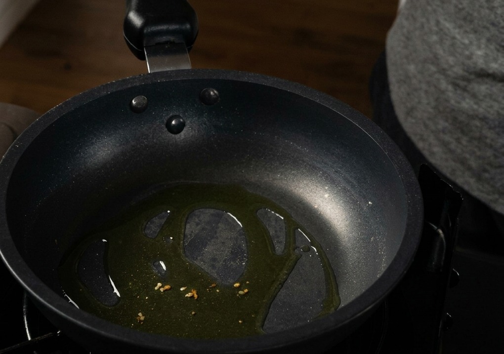 Pezzetti di aglio tritato che sfrigolano nellolio doliva sul fondo di una padella antiaderente nera.