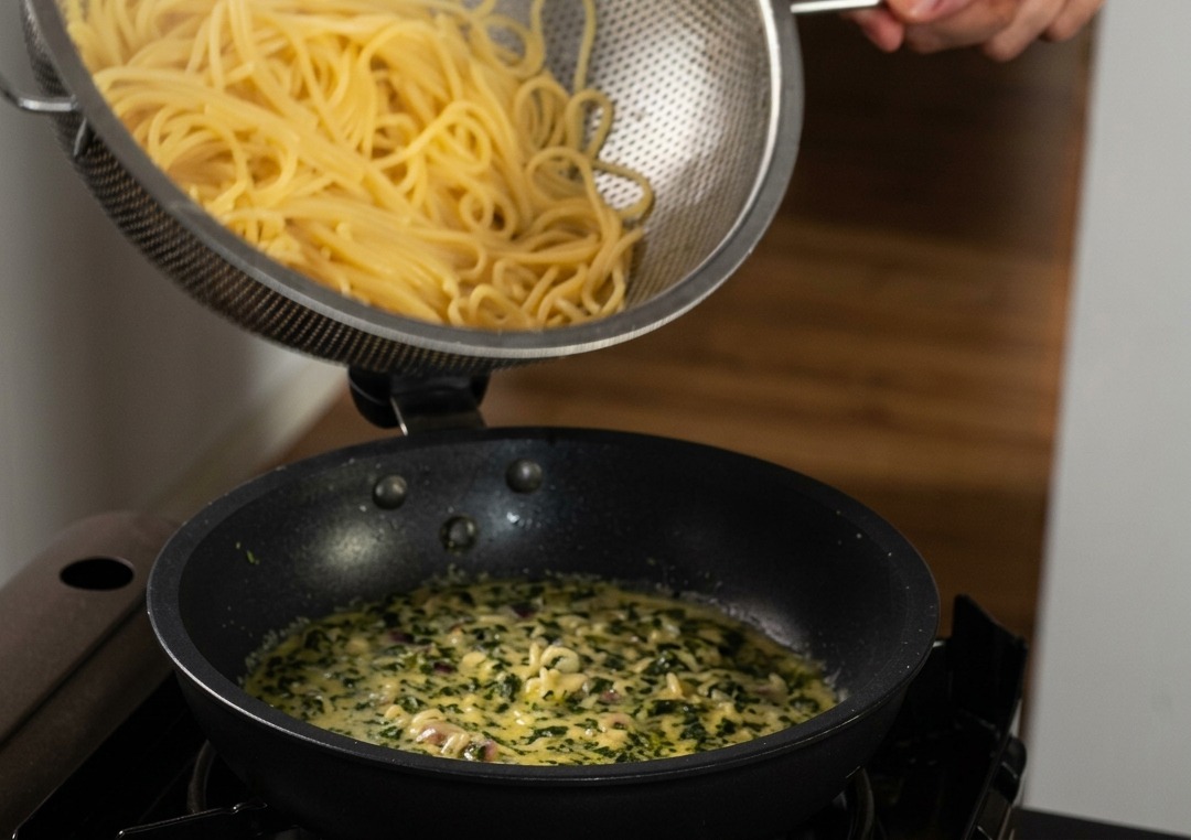 Spaghetti cotti versati da uno scolapasta in acciaio inossidabile dentro una padella colma di salsa cremosa e basilico verde.