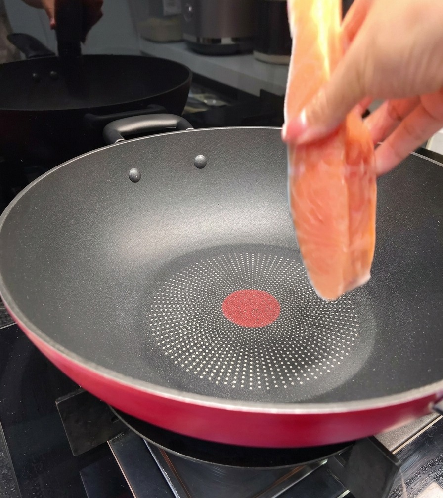 Una mano che adagia un filetto di salmone fresco in una grande padella antiaderente nera sul piano cottura.