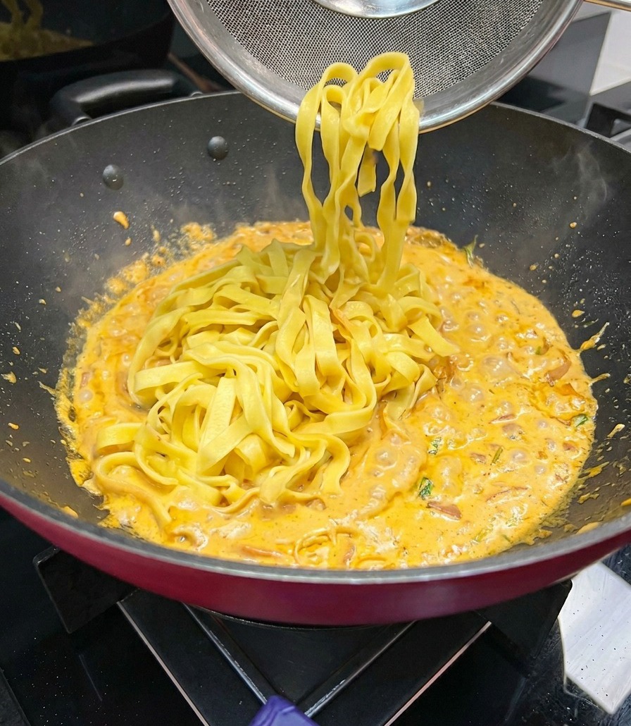 Fettuccine larghe sollevate con un mestolo a ragno e aggiunte a una padella con salsa cremosa arancione in ebollizione.