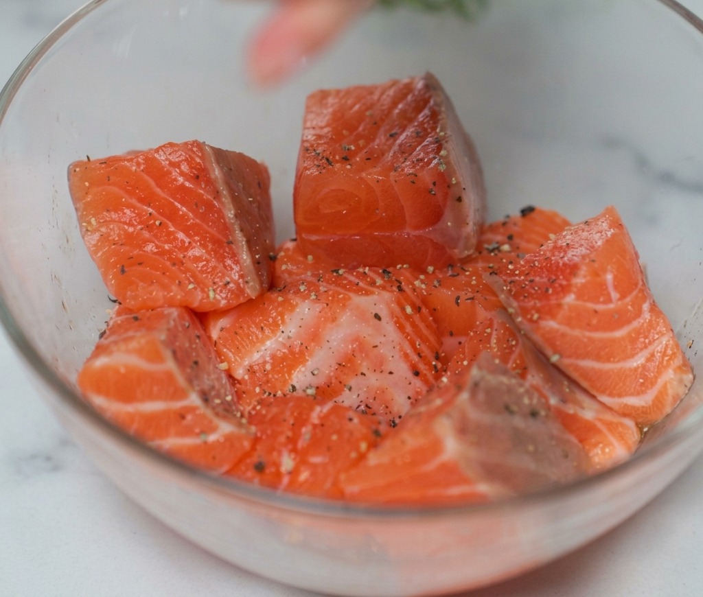 Cubetti di salmone crudo in una ciotola di vetro mentre vengono conditi con sale e pepe nero da un macinino.