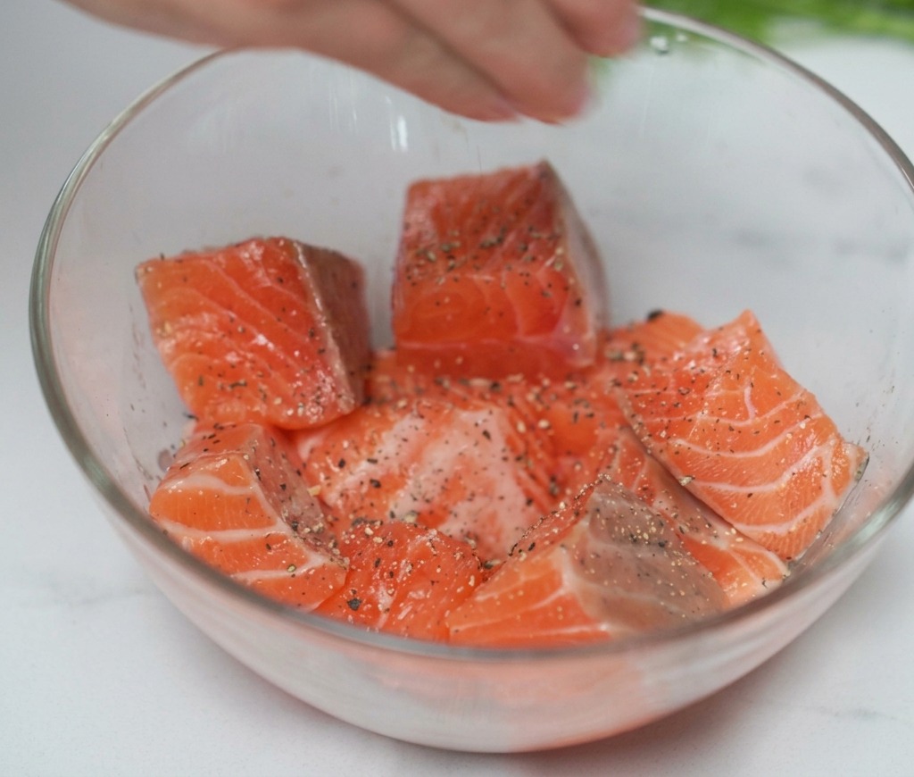 Una mano che spreme succo di limone fresco sui cubetti di salmone conditi in una ciotola trasparente.