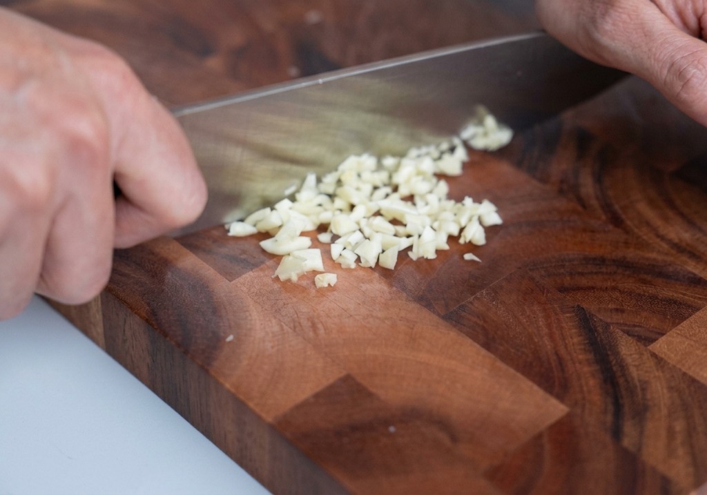 Aglio tritato finemente su un tagliere di legno con un coltello da chef accanto.