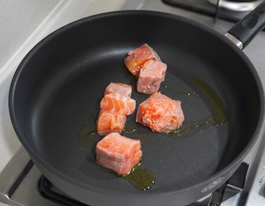 Cubetti di salmone che cuociono in una padella antiaderente nera con olio doliva su un fornello a gas.